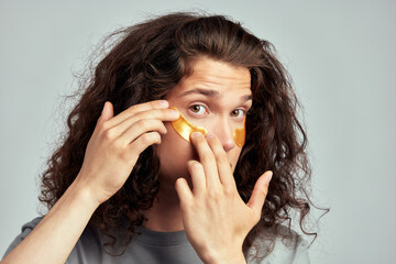 Young handsome long-haired guy applying under eye hydrogel patches on his face. Caucasian brown-haired millennial man practices skin care routine to keep healthy and youthful looking. Studio portrait.