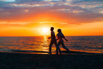 Silhouette of a young couple in love together holding hands and running along the seashore at sunset. Romance, relationships, valentines day concept