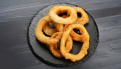 delicious golden battered, breaded and deep fried crispy onion rings served on round black stone tray on black wooden table, view from above, close-up
