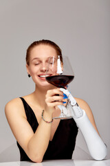 Young smiling girl with disability wearing sensory bionic prosthetic arm seats at the table. Beautiful woman holds a glass of red wine with her artificial robotic hand. Full life after limb loss.