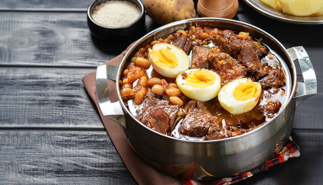 close-up of Traditional Jewish Cholent (Hamin) - main dish for the Shabbat meal slow cooked beef with potato, beans and brown eggs in a metal casserole on a black wooden table