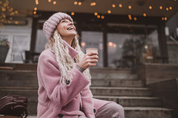 fashionable mature woman in pink knitted beanie hat and stylish coat sits outside in cafe and drinks coffee from disposable paper cup. Middle aged woman positive and smiling with pigtails in the city 