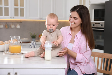 Mother and her cute little baby with feeding bottle of milk in kitchen, space for text