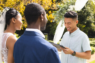 Happy diverse male officiant using tablet, with bride and groom at wedding ceremony in sunny garden