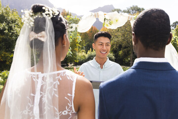 Happy diverse male officiant using tablet, with bride and groom at wedding ceremony in sunny garden