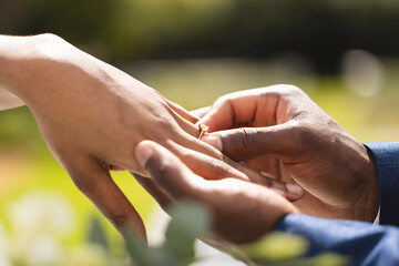 Hands of african american bride and groom placing ring on finger at wedding ceremony in sunny garden