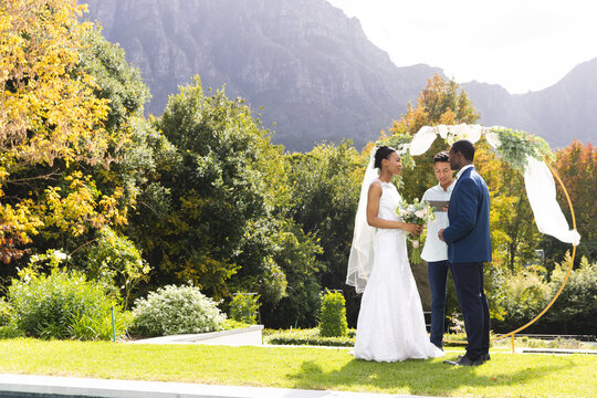 Happy diverse male officiant, bride and groom at outdoor wedding in sunny garden, copy space