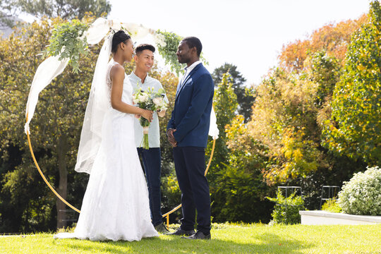 Happy diverse male officiant, bride and groom at outdoor wedding in sunny garden, copy space - Powered by Adobe