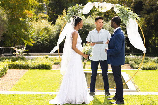 Happy diverse male officiant, bride and groom at outdoor wedding in sunny garden, copy space