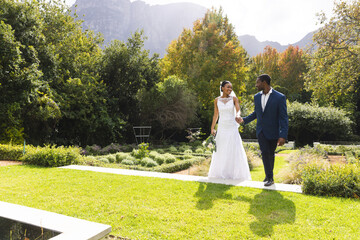 Happy african american bride and groom holding hands and walking in sunny garden, copy space