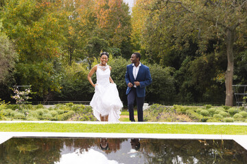 Happy african american bride and groom on wedding day walking together in garden, copy space
