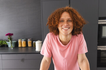 Portrait of happy biracial man with curly red hair leaning on counter in modern kitchen