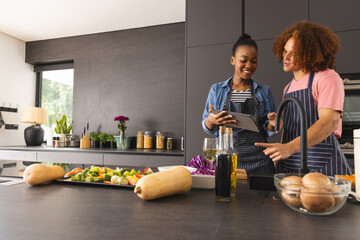Happy diverse couple in aprons preparing meal, using tablet in kitchen, copy space