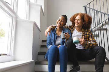 Happy diverse couple sitting on stairs at home, drinking coffee, embracing and looking out of window