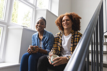 Portrait of happy diverse couple sitting on stairs at home drinking coffee