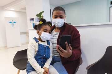 African american mother and daughter wearing face masks using smartphone in waiting room at hospital
