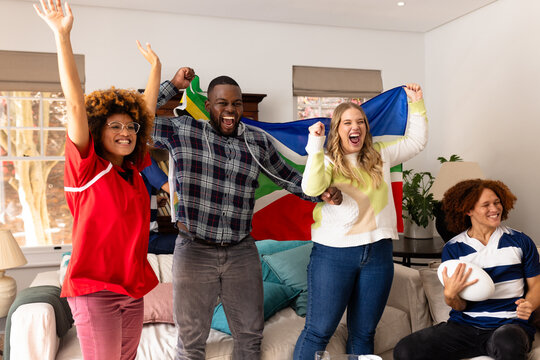 Happy group of diverse friends with rugby ball and flag of south africa supporting at home