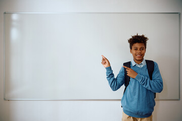 Happy African American student pointing at copy space on whiteboard in classroom while looking at...