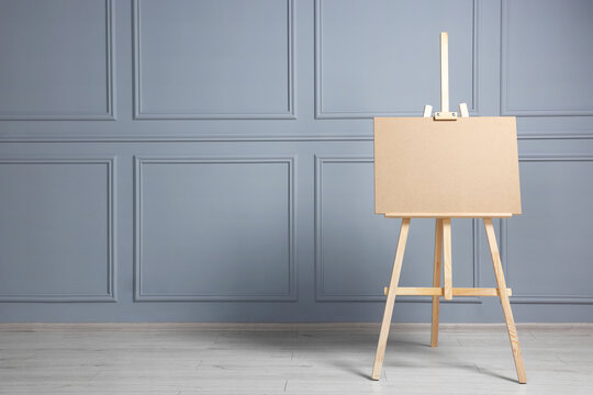 Wooden easel with blank board near grey wall indoors. Space for text