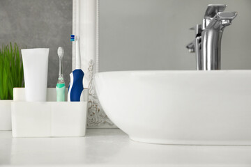 Fototapeta premium Electric toothbrushes and tube of paste near vessel sink on countertop in bathroom, closeup