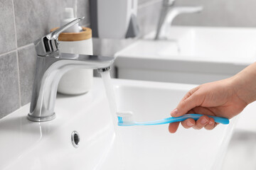 Woman washing plastic toothbrush under flowing water from faucet in bathroom, closeup