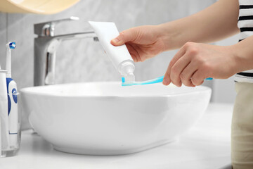 Woman squeezing toothpaste from tube onto toothbrush near sink in bathroom, closeup