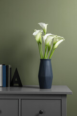 Beautiful calla lily flowers in vase and books on grey chest of drawers near olive wall