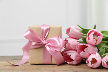 Beautiful gift box with bow and pink tulips on wooden table, closeup