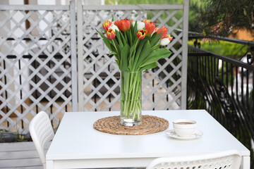 Fototapeta na wymiar Beautiful colorful tulips in glass vase and cup of drink on white table at balcony