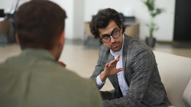 Successful broker advisor wearing eyeglasses and business suit consulting talking to unrecognizable man client sitting at desk. Confident male insurer consulting consumer about insurance benefits.