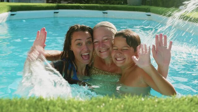 Grandmother, adult daughter, and grandson relaxing in the swimming pool on summer vacation together smiling and waving looking at camera.