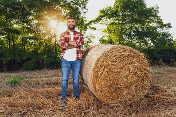 Naklejka na ściany i meble Farmer is standing beside bales of hay. He is satisfied after successful harvesting.