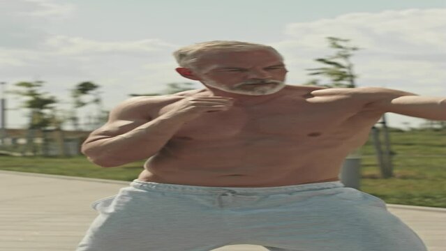 Vertical shot of shirtless senior man with muscular torso practicing boxing with assistance of personal trainer outdoors