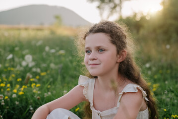 Fototapeta na wymiar Portrait of happy preteen girl sitting on green grass outdoor, enjoying beautiful summer nature during warm sunset or sunrise. People emotions, travel, adventure, childhood and wanderlust concept