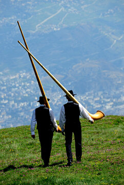 Nendaz, Conthey District, 
Valais, Wallis, Switzerland, Europe - two men in traditional black vests, trousers and hats walking with alphorn on their arms, Swiss Alps, Nendaz village seen in valley 