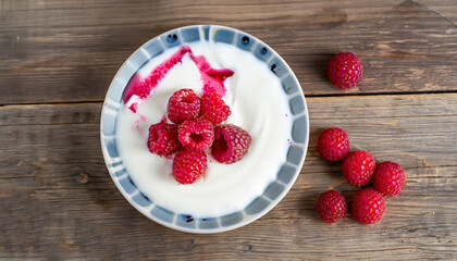 fresh greek yogurt with colored stains of berry juice and whole raspberry, dewberry on plate on old wooden table, view from above