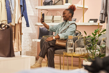 Full length portrait of smiling black young woman clothing boutique and using smartphone, copy space