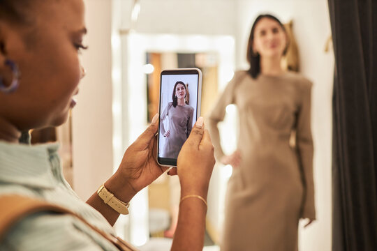 Closeup of young woman taking photos of friend trying on clothes in dressing room at boutique