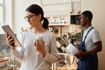 Side view portrait of woman using digital tablet at pharmaceutical factory and holding bottle of pills