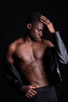 Young black man in jacket touching head and showing abs in light