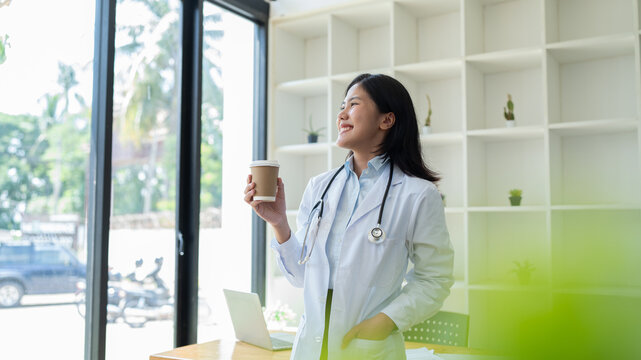 Young Asian Woman Doctor With Coffee Cup In Medical Office In Hospital. Resting Taking Break Standing Looking Out Window In Clinic