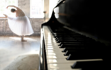 A young ballerina in a white tutu rehearses a dance against the background of a grand piano in a...