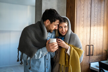 Happy young couple with hot drinks cuddling under warm blanket at home