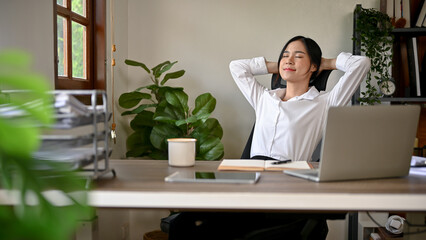 A happy and relaxed Asian businesswoman is leaning on her chair, putting hands behind her head
