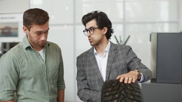 Portrait of professional salesman wearing business suit talking about characteristic of tires product to customer male came to look at assortment represented in auto service shop, slow motion.