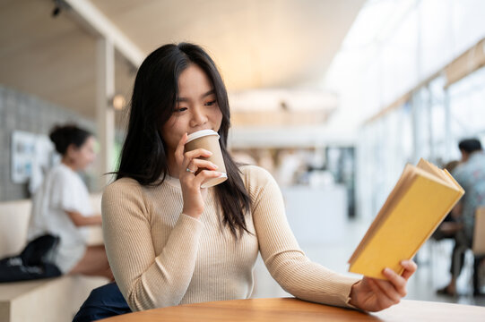 A Beautiful Asian Woman Is Sitting In A Coffee Shop, Reading A Book, And Enjoying Her Coffee.