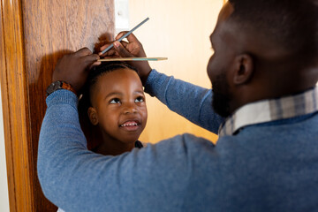 Happy african american father measuring son and marking height on door frame at home