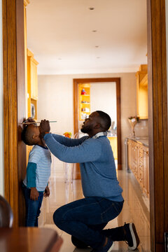 Happy african american father measuring son and marking height on door frame at home, copy space