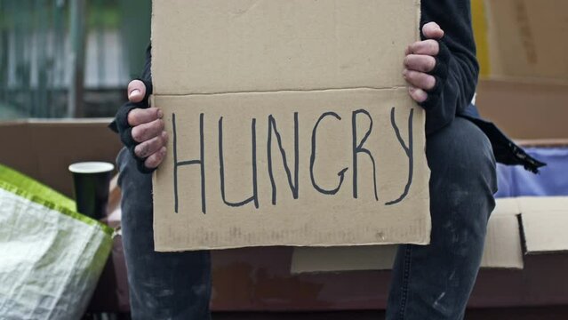 An untidy man is sitting with a cup of some drink and holding a handwritten HUNGRY placard.
