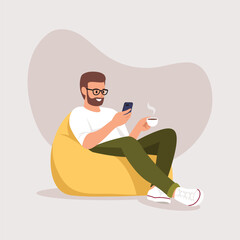 a man sits on an armchair (bean bag) with a phone and a cup of coffee